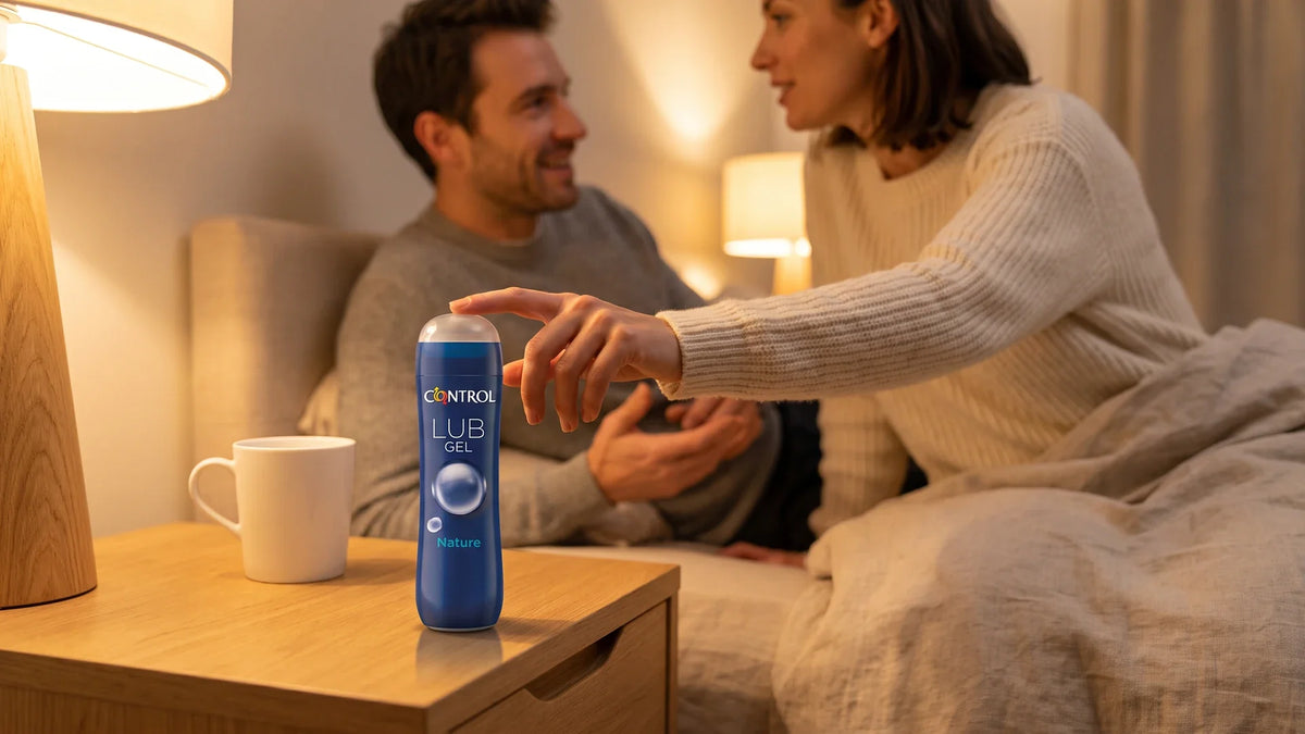 Femme en pull crème effleurant le bouchon du lubrifiant Control Lub Gel Nature posé sur une table de chevet, son compagnon souriant en arrière-plan