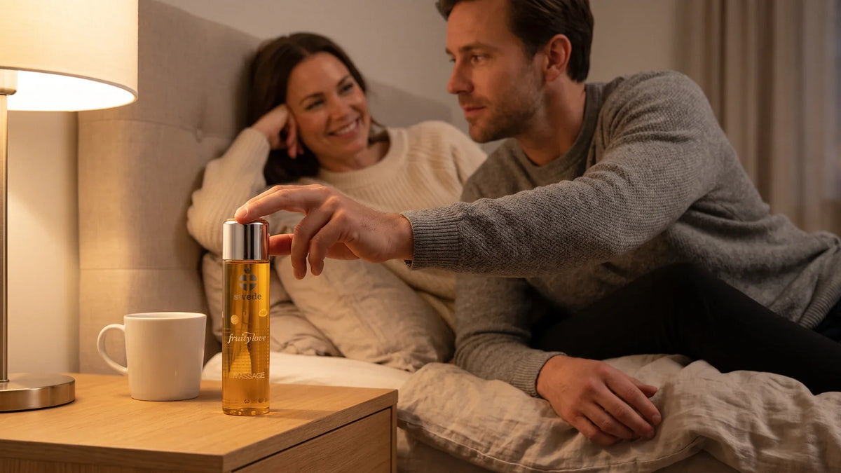 Homme assis au bord du lit touchant un flacon d’huile de massage Swede sur le chevet, sa partenaire souriante derrière lui